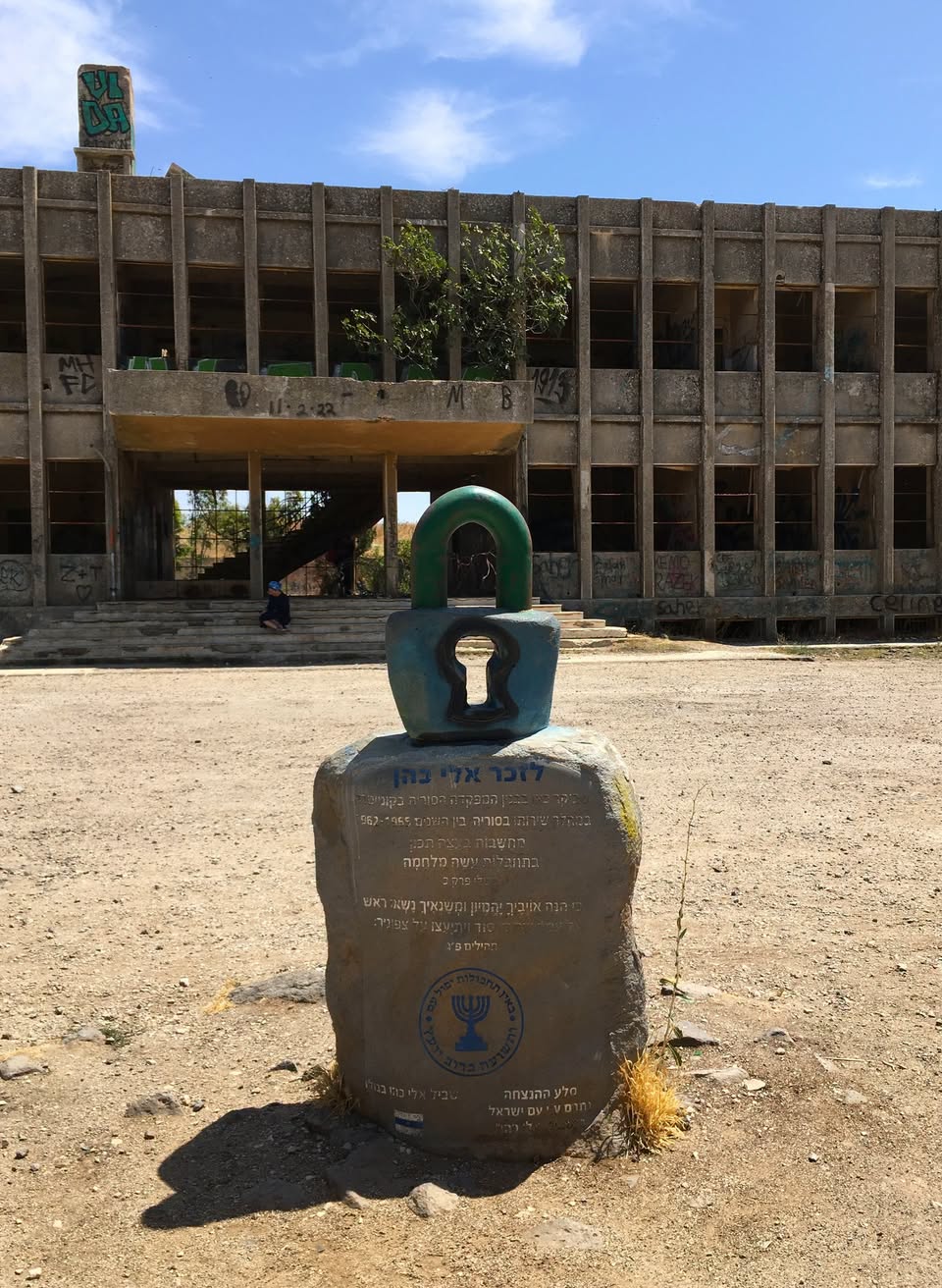 A memorial stone to Eli Cohen topped by a padlock sculpture before an abandoned Syrian army building on the Golan Heights